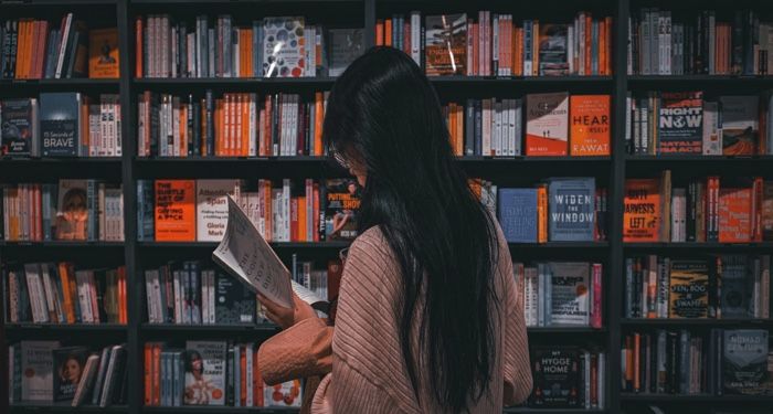 tan skinned person with long black straight hair reading a book in front of a book stores shelves.jpg.optimal