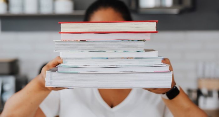 tan skinned person is holding a stack of books in front of them obscuring their face.jpg.optimal